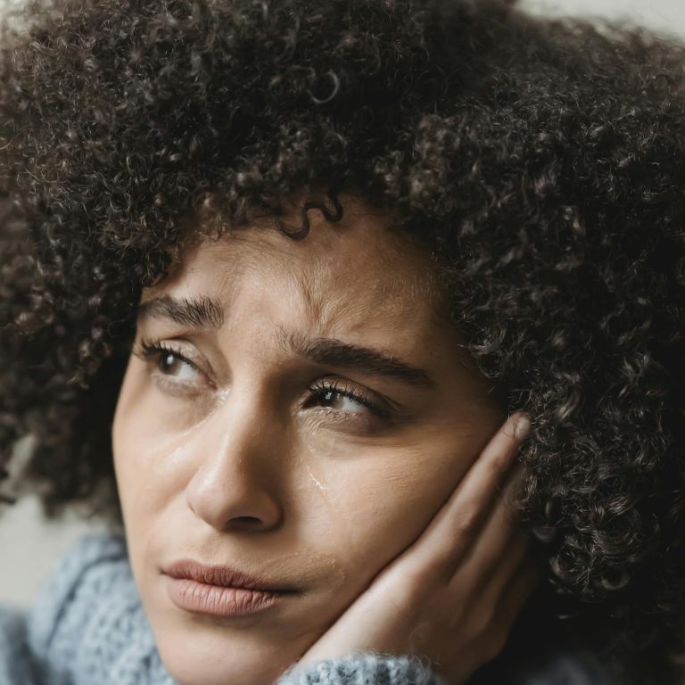 A thoughtful woman with curly hair leans on her hand, wearing a cozy sweater.